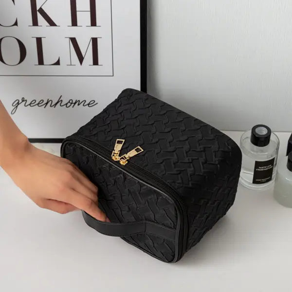 Black woven cosmetic bag with gold zipper on a white surface, with a hand reaching towards it.
