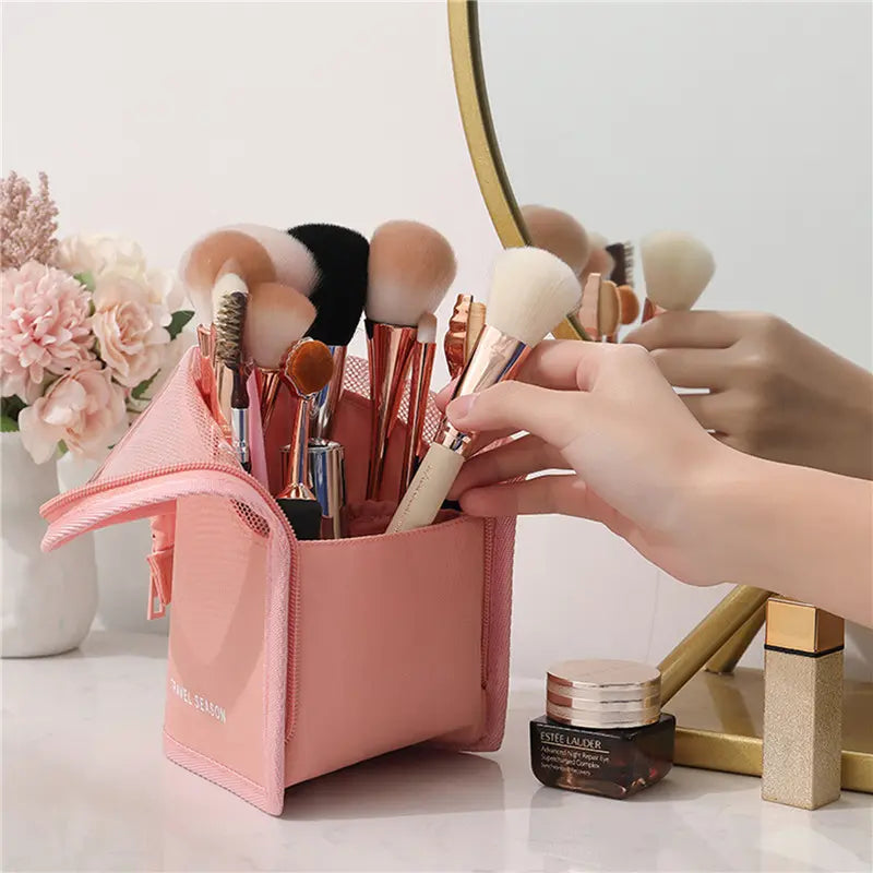 Makeup brushes in a pink holder with a hand selecting a brush, set against a blurred background of flowers and a mirror.