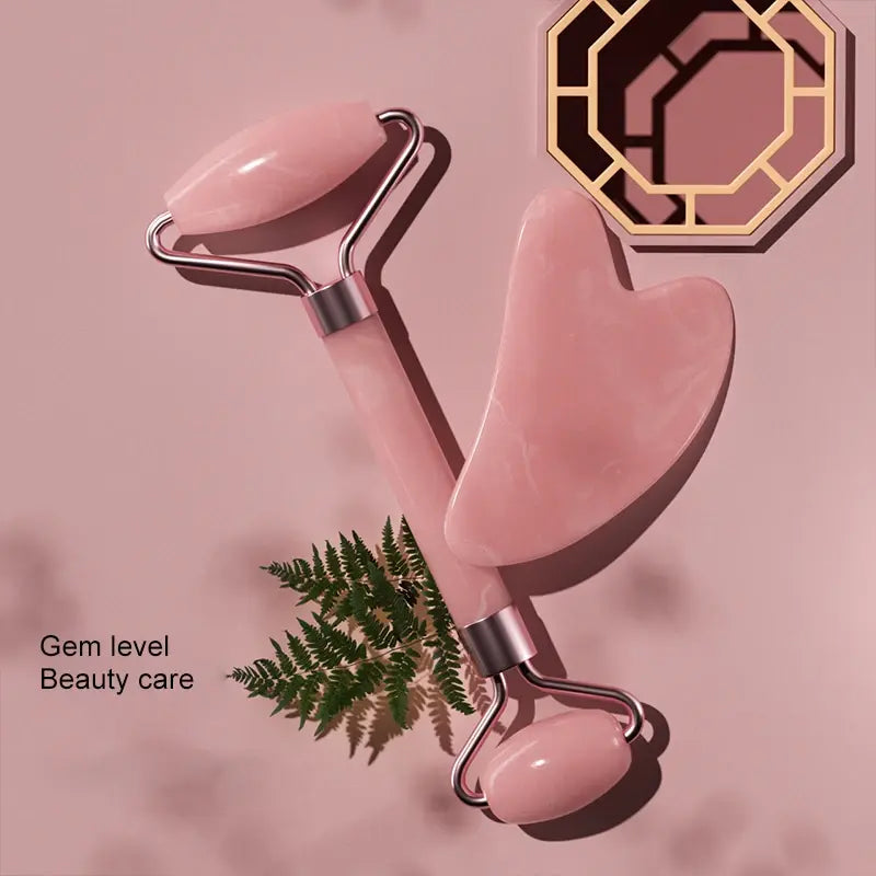 Pink facial roller and gua sha tool on a pink background with fern leaves.